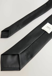 Cravate noire à motifs avec une surface texturée et une doublure unie. Présente une étiquette de "NEXT Tailoring", positionnée à une extrémité.