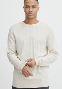 Suéter de punto color crema con cuello redondo, mangas largas, puños y dobladillo acanalados. Textura suave con un sutil patrón jaspeado.