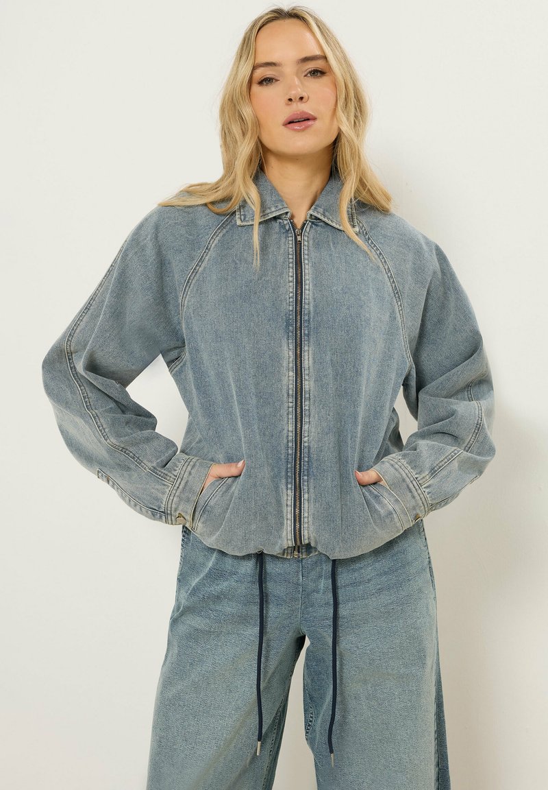 Blondynka w jasnoniebieskiej kurtce dżinsowej na suwak z kieszeniami i dopasowanych spodniach dżinsowych, stojąca na jednolitym tle.