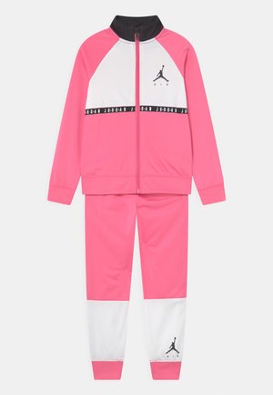 Różowo-biały dres Jordan z czarnym kołnierzem, kurtka na pełny zamek, logo na piersi i spodniach oraz pas z napisem „Jordan” na wysokości klatki piersiowej.