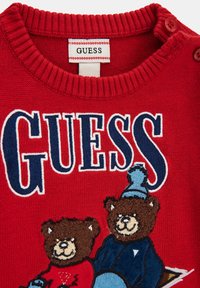 Czerwony sweter z dzianiny z dwoma haftowanymi brązowymi niedźwiedziami, z których jeden ma na sobie niebieski kapelusz. Marka "GUESS" jest wyraźnie widoczna w granatowym i białym napisie.