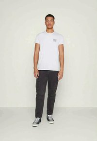 T-shirt branca de algodão com pequeno texto preto no peito, combinada com calças cargo cinza-escuras e ténis pretos de cano alto.