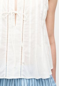 Λευκό αμάνικο μπλουζάκι με κάθετες διάτρητες ρίγες, λεπτομέρεια με κορδόνι μπροστά, συνδυασμένο με μπλε και μαύρο παντελόνι με κάθετες ρίγες.