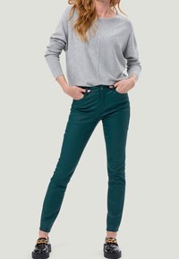 Haut gris à manches longues avec une coupe décontractée, associé à un pantalon vert foncé ajusté. Le modèle porte des chaussures noires avec de fortes accents dorés.
