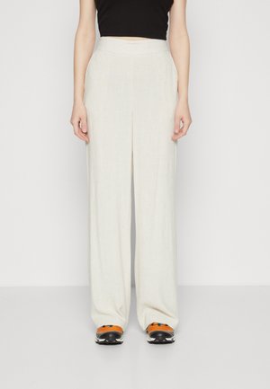 Femme portant un pantalon beige clair à taille haute et jambes larges, associé à des baskets orange et noires ainsi qu'un top noir court, se tenant devant un mur blanc.