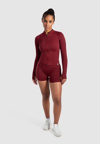Bordeauxkleurige sportset bestaande uit een lange mouw zip-up top en bijpassende shorts, gemaakt van rekbare stof met een gladde textuur.