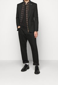 Giacca nera con rever a punta sopra una camicia a quadri. Pantaloni neri e scarpe nere. In generale, un look formale e su misura.