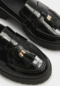Mocassini neri in pelle verniciata con dettaglio a nappa, accenti in hardware dorato e suole in gomma spesse. Presentano una finitura liscia e lucida.