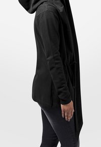 Chaqueta con capucha negra que presenta un largo frontal con una silueta abierta, textura suave y bolsillos laterales, combinada con jeans oscuros.