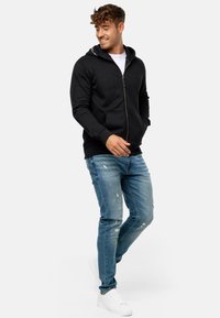 Sorte zip-up hættetrøje med snoretræk i hætten, frontlommer og ribstrikkede manchetter, parret med slidte blå jeans og hvide sneakers.