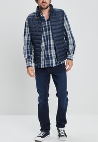 Chaleco acolchado azul marino con cuello alto sobre una camisa a cuadros azul y blanca, combinado con jeans oscuros y zapatillas azul marino. Textura suave y diseño ligero.