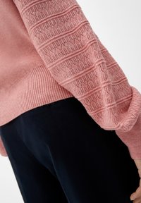 Gros plan sur une personne portant un pull en maille rose texturé avec une manche à motif détaillé et un pantalon foncé.