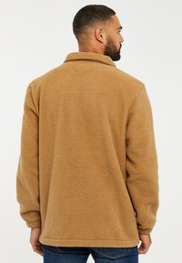 Chaqueta de forro polar color beige con un corte relajado, cuello alto y puños acanalados. Tejido texturizado con un aspecto suave y acogedor, vista desde la parte de atrás.
