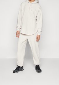 Persoon in een off-white hoodie en bijpassende broek met zwart-grijze Nike sneakers, staand op een effen lichte achtergrond.