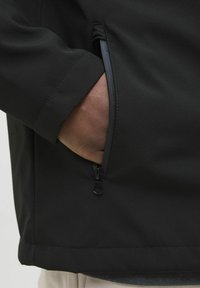 Sort softshell-jakke med en lynlåslomme på siden, tekstureret stof og en glat, holdbar finish. Søm detaljer ved kanten.