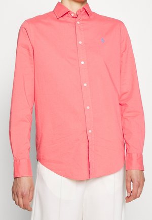 Homme portant une chemise corail à manches longues avec des boutons et un petit logo bleu brodé sur le côté gauche de la poitrine, ainsi qu'un pantalon blanc.