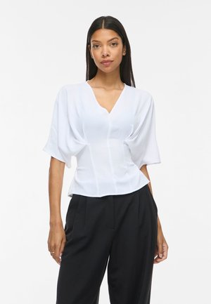 Femme portant un chemisier blanc à col en V avec des manches larges et un pantalon noir taille haute, debout sur un fond blanc uni.