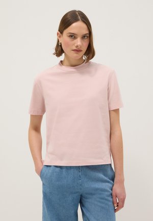 Joven con cabello corto y castaño, lleva una blusa de manga corta de color rosa claro y pantalones de mezclilla azules, de pie con una mano en el bolsillo.