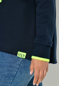 Hand ruhend an der Seite, getragen wird ein dunkelblaues Langarmshirt mit neongrünem Besatz und Jeans, vor neutralem Hintergrund.