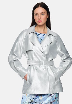 Woman with straight dark hair wearing a shiny light silver belted trench coat over a blue floral dress, standing with hands in pockets.