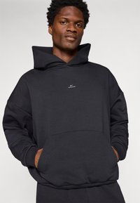Sudadera negra con una capucha grande, bolsillo frontal y puños acanalados. Presenta un sutil logo blanco en el pecho. Fabricada con una tela suave y cómoda.