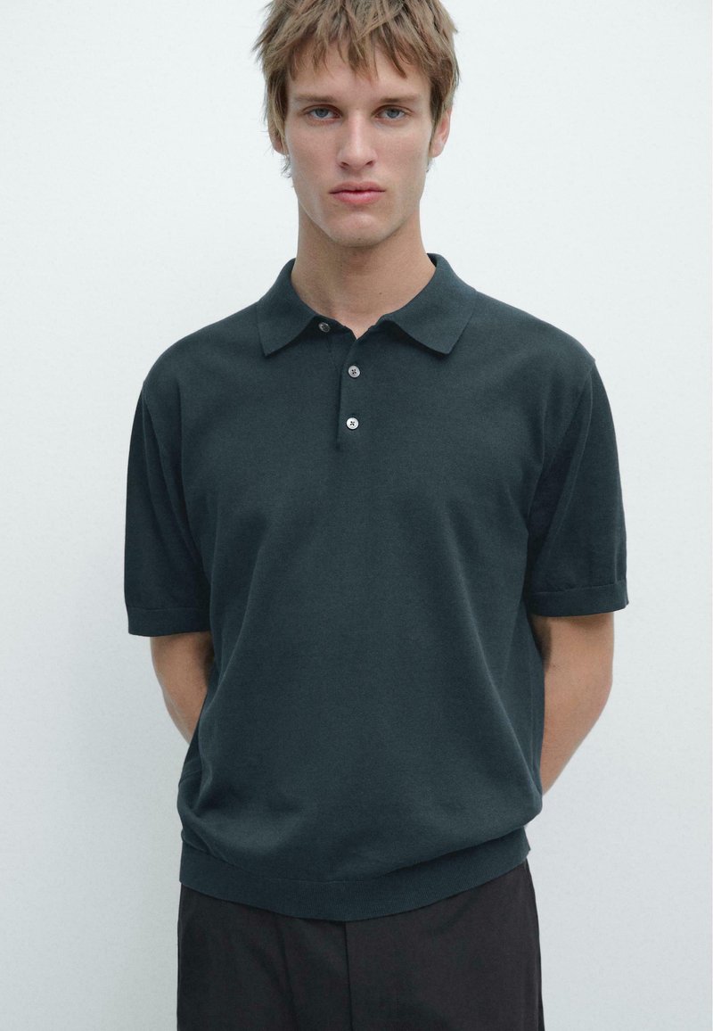 Jeune homme aux cheveux courts brun clair portant un polo manches courtes vert foncé et un pantalon sombre, debout devant un fond uni.