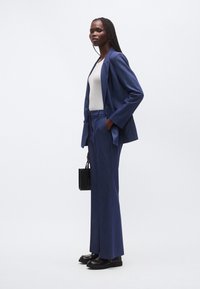 Tmavomodrý na mieru šitý oblek s priliehavým sako a nohavicami s širokými nohavicami, kombinovaný so svetlosivým topom a čiernymi mokasínmi. Doplnky zahŕňajú malú čiernu tašku.