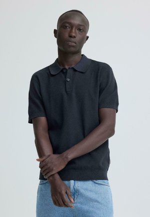 Jonge man met kort haar, gekleed in een donkerblauw poloshirt en lichtblauwe jeans, staand tegen een effen lichte achtergrond, met gekruiste armen.