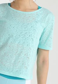 Ljus turkos kortärmad topp med ett strukturerat mönster, rund halsringning och en avslappnad passform. Underlag i matchande färg är synligt.
