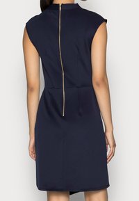 Kvinna klädd i en ärmlös marinblå klänning med en synlig gulddragkedja som löper vertikalt ner längs ryggen.