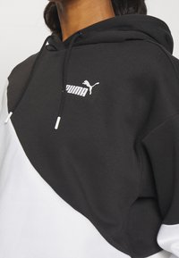 Svart och vit huvtröja med en diagonalt mönster, med en Puma-logotyp i vitt och snoddar med silverfärgad spets. Mjukt material.