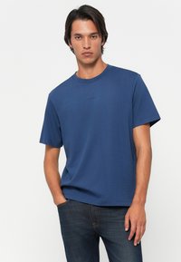 Young man with dark hair wears a plain blue T-shirt and dark jeans, standing with one hand in pocket against a plain white background.
