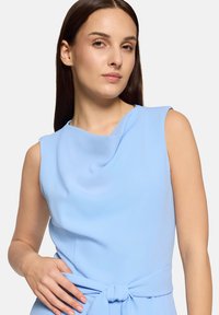 Robe bleu clair sans manches avec un col drapé, un détail de ceinture nouée, et une texture de tissu lisse, mettant en valeur une silhouette cintrée.