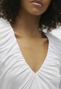 Abito bianco con scollo a V profondo e dettagli di tessuto arricciato. Il materiale è liscio, mettendo in risalto la texture del capo intorno allo scollo.