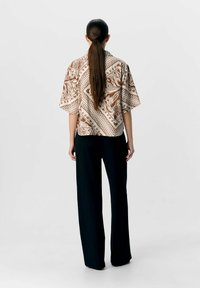 Camicia corta a maniche corte di colore marrone e beige con un motivo ricamato, abbinata a pantaloni neri larghi. Il tessuto presenta dettagli floreali intricati.