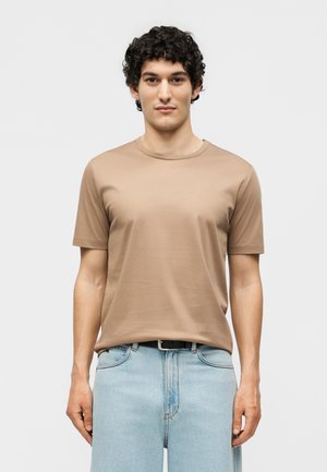 Jeune homme aux cheveux bouclés portant un t-shirt beige uni à manches courtes, rentré dans un jean bleu clair avec une ceinture noire, debout devant un fond blanc.