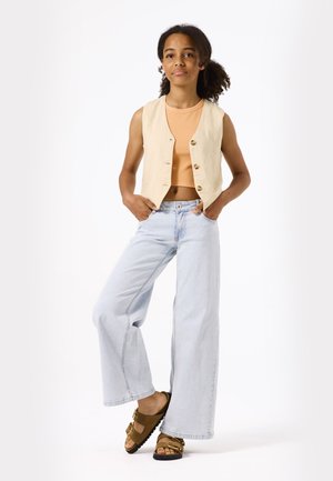 Beige linnen vest met knoopsluiting, gecombineerd met een lichtoranje crop top en lichtblauwe wijde jeans. Bruine sandalen met gouden hardwareaccenten.