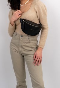 Schwarze Leder-Umhängetasche mit geschwungener Form, goldenem Reißverschluss und Markenlogo. Die Person trägt einen beigen Pullover und hellfarbige Hose.