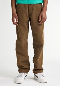Pantaloni in corduroy marrone con una texture a coste, vestibilità dritta e tasche laterali, abbinati a sneakers bianche con accenti neri.
