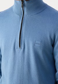 Pull léger bleu clair à fermeture éclair avec un col montant, fabriqué en tissu doux, doté d'un écusson carré avec logo sur la poitrine gauche. Motif en tricot texturé.