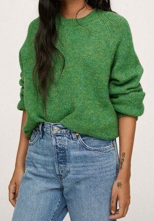 Kvinde iført en grøn strikket sweater med opkørte ærmer stoppet ned i højtaljede blå jeans, der viser tatoveringer på hendes højre underarm og hånd.