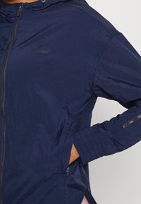 Námořnická modrá lehká bunda vyrobená z texturované látky se zipem, kapucí a bočními kapsami. Má žebrované manžety a detail s logem.