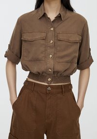 Femme portant une chemise marron courte à boutons avec les manches retroussées et un pantalon marron taille haute assorti, avec poches et fermeture à boutons.