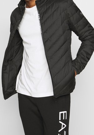 Homme portant une veste matelassée noire sur un t-shirt blanc avec un pantalon noir comportant un logo EA7 vertical blanc sur la jambe gauche.