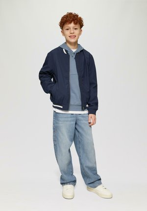 Marine bomberjack over een lichtblauwe hoodie, gecombineerd met losse lichtblauwe jeans en witte sneakers; gekruld rood haar; casual outfit.