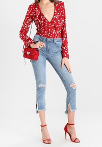 Blusa larga de flores rojas con mangas largas y escote en V profundo, combinada con jeans rasgados de un azul claro y tacones rojos con tiras. Incluye un pequeño bolso rojo.
