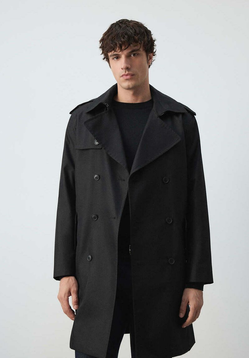 Homme aux cheveux courts et bouclés portant un manteau noir croisé et une chemise noire sur un fond clair uni.