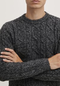 Tamsiai pilkas megztinis su kabelių raštu, apvaliu kaklu ir tekstūruota medžiaga. Rankovės baigiasi elastiniais raukiniais.