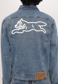 Giubbotto di jeans azzurro chiaro con una grafica bianca di un gatto che salta sulla schiena, dotato di una toppa in pelle beige con il marchio in vita.