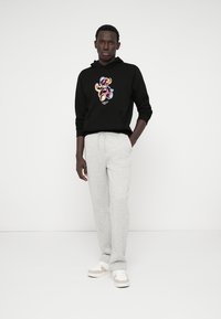 Zwarte hoodie met kleurrijke teddybeer graphic, gecombineerd met lichtgrijze sweatpants. Model staat tegen een effen achtergrond. Witte sneakers maken de look compleet.